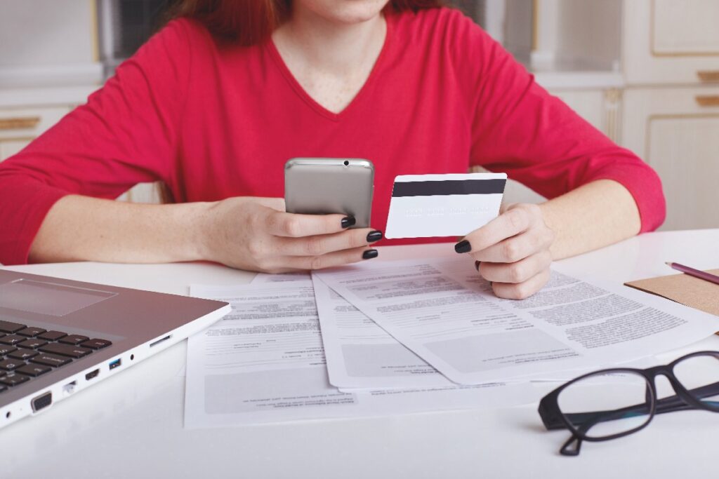 Torso de mujer con jersey rojo, con una tarjeta de crédito en una mano y un móvil en la otra, sobre mesa con documentos, ordenador portátil y gafas